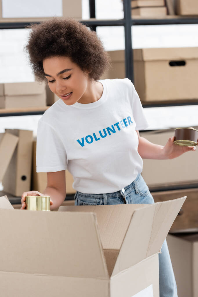 sorridente volontario afroamericano che tiene cibo in scatola vicino alla scatola in un magazzino di beneficenza - Foto, immagini