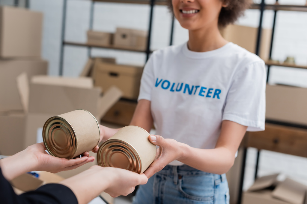 vista recortada de sonriente afroamericano voluntario repartiendo comida enlatada en el centro de caridad - Foto, Imagen