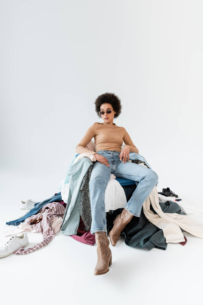 afro-amerikai nő divatos napszemüveg ül közelében különböző ruhák szürke háttér - Fotó, kép