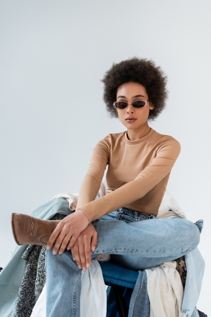 curly african american woman in trendy sunglasses sitting near clothes on grey background - Fotó, kép