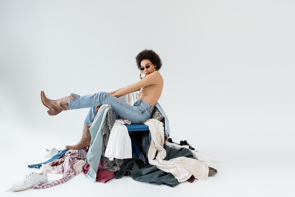 stylish african american woman in sunglasses and cowboy boots posing near pile of clothing on grey background - Valokuva, kuva