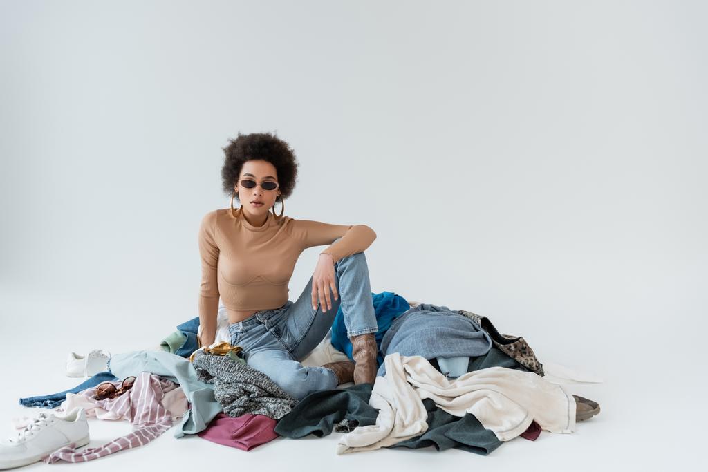 fashionable african american woman in sunglasses near pile of garments on grey background - Photo, Image