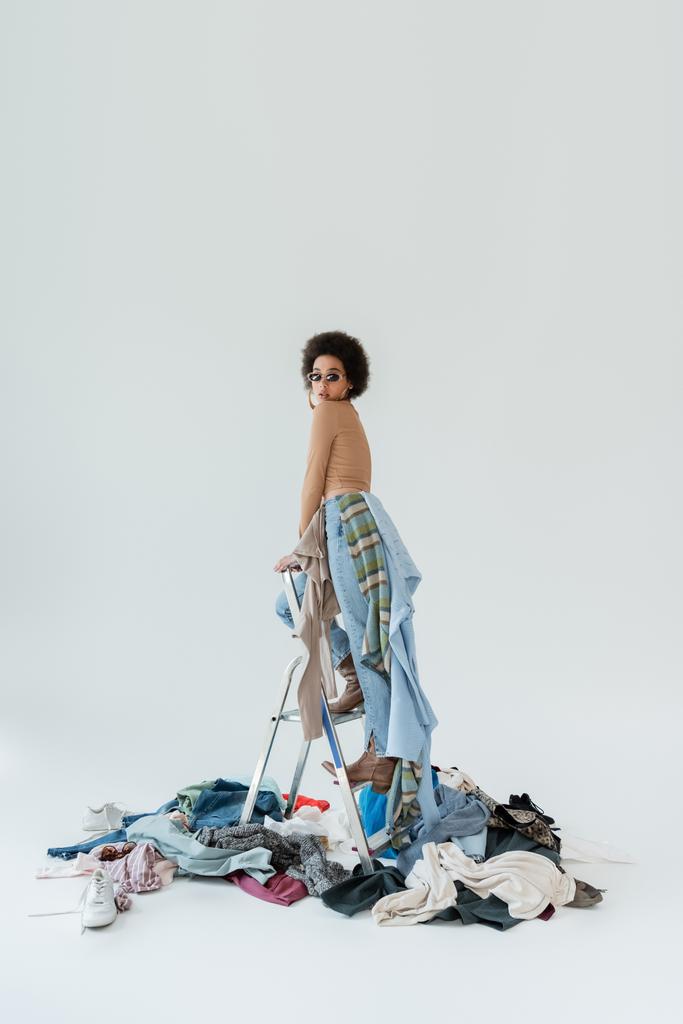 trendy african american woman standing on ladder near pile of clothes on grey background - Photo, Image