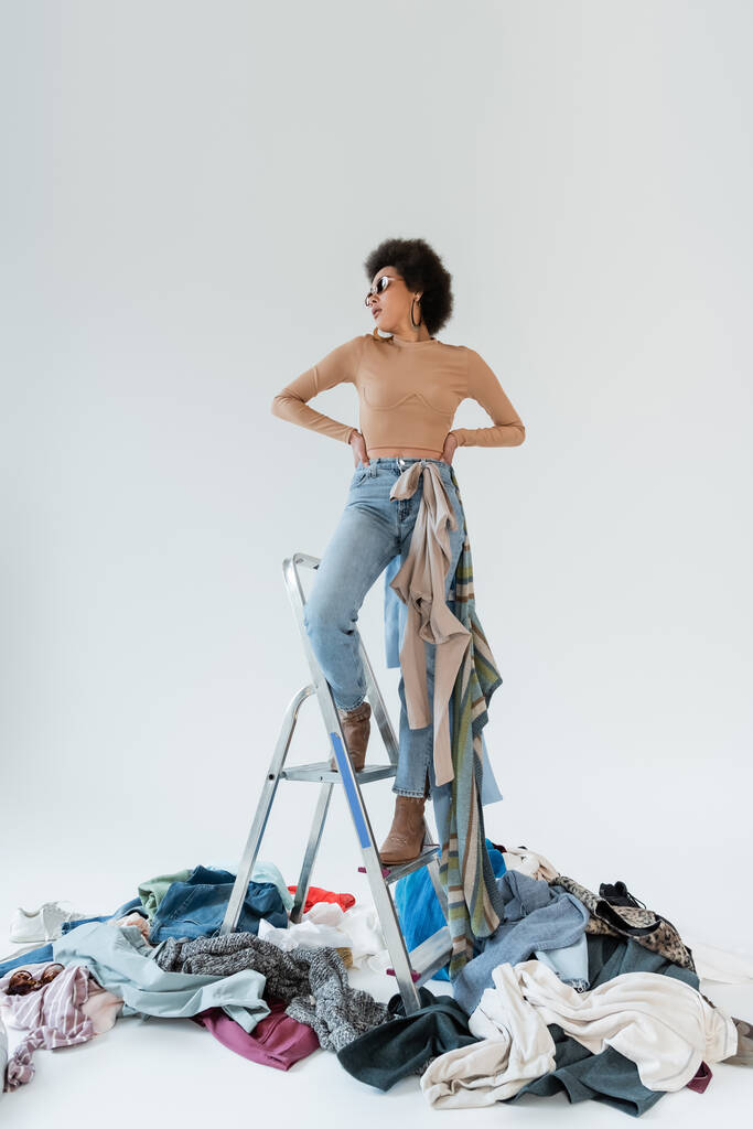 african american woman with hands on waist standing on ladder near mess of garments on grey background - Foto, imagen