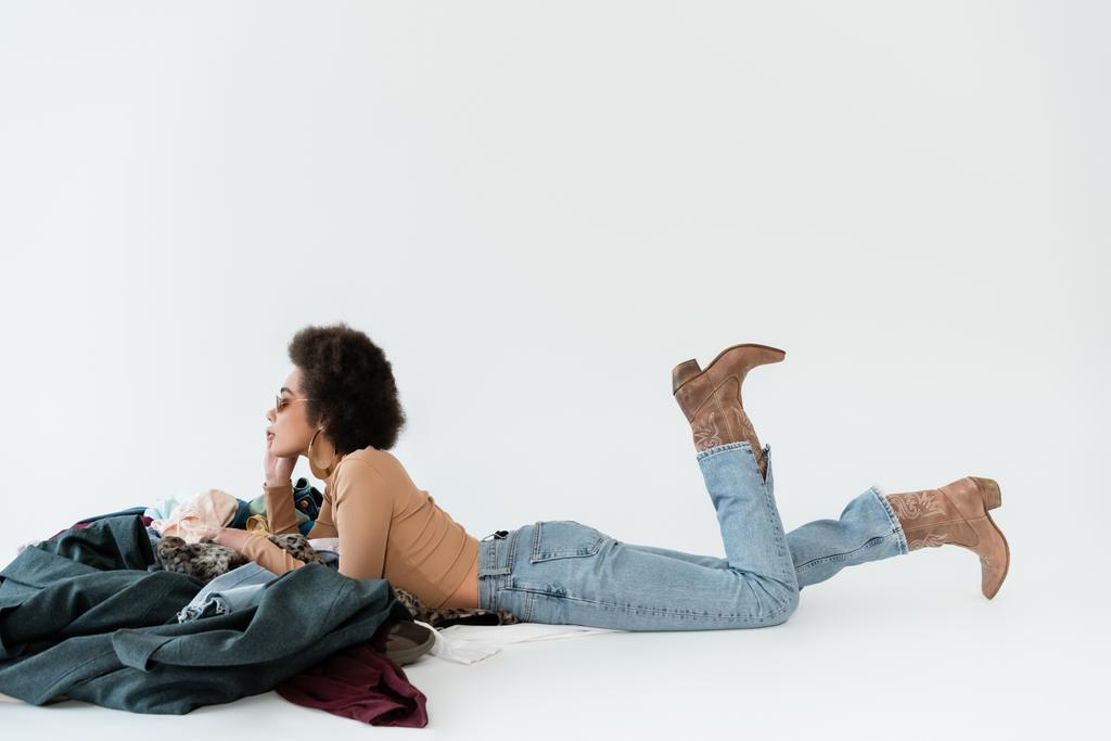 vista lateral da mulher afro-americana em jeans e botas de cowboy deitado perto de pilha de roupas em fundo cinza - Foto, Imagem
