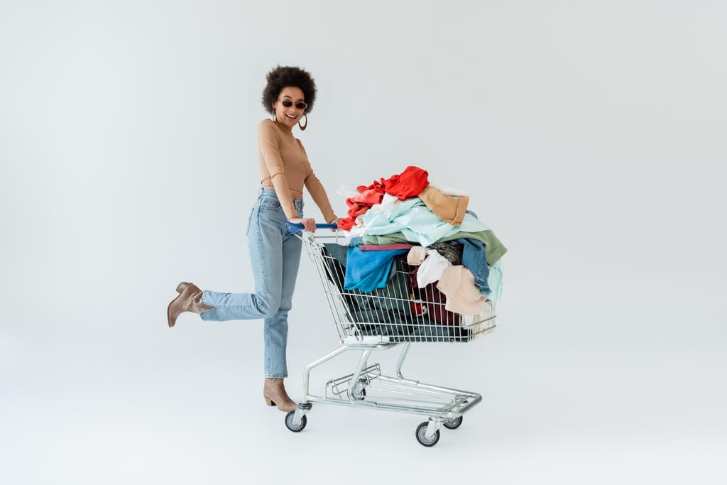 longitud completa de la mujer afroamericana feliz cerca del carrito de compras lleno de ropa sobre fondo gris - Foto, Imagen