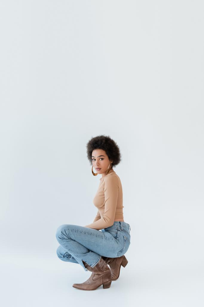 donna afroamericana alla moda in camicia a maniche lunghe beige e jeans in posa su sfondo grigio - Foto, immagini