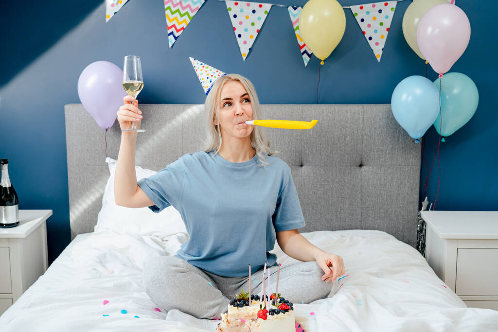 Happy emotional woman in pajama and party cap blowing in birthday pipe while sitting on the bed with festive cake and champagne glass. Time for yourself Celebration at home. Happy birthday concept - Foto, Imagem