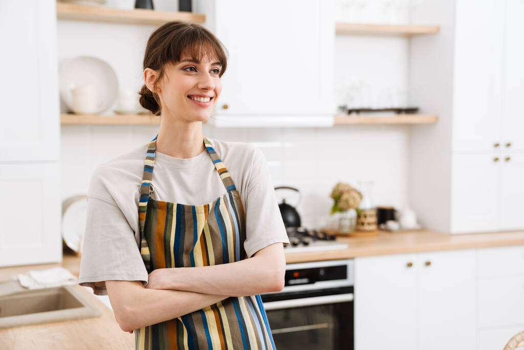 Λευκή νεαρή γυναίκα που φοράει ποδιά χαμογελώντας ενώ στέκεται στην κουζίνα στο σπίτι - Φωτογραφία, εικόνα