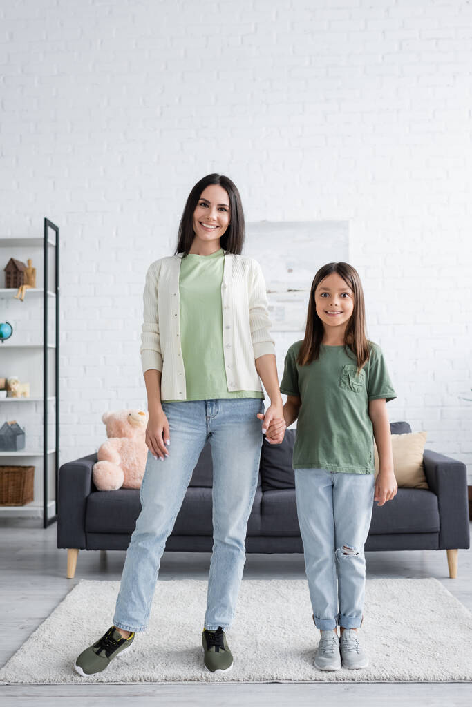 full length of cheerful woman and girl standing and holding hands in modern living room  - Valokuva, kuva