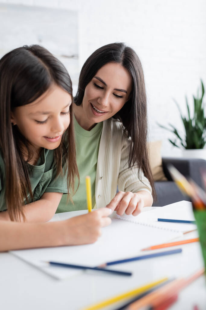 cheerful babysitter looking at paper while girl drawing with yellow pencil  - Foto, Imagem
