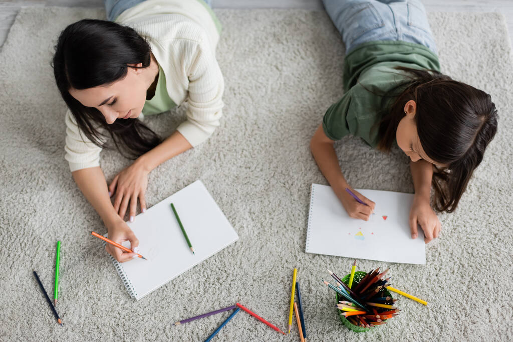 top view of nanny and girl drawing on papers while lying on carpet in living room  - Fotoğraf, Görsel