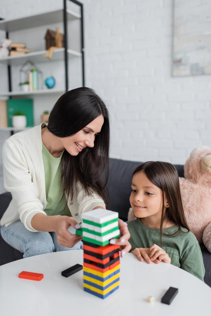 happy babysitter playing wood blocks game with pleased girl in living room - Foto, Imagem