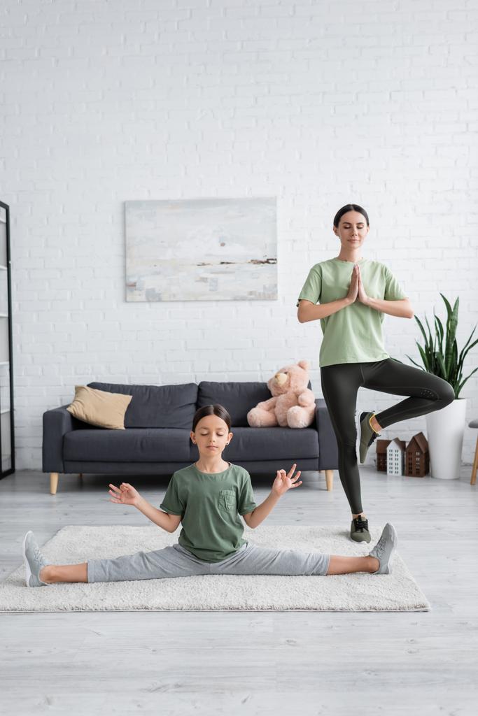 longitud completa de niñera y chica con los ojos cerrados meditando en yoga posa en sala de estar - Foto, imagen