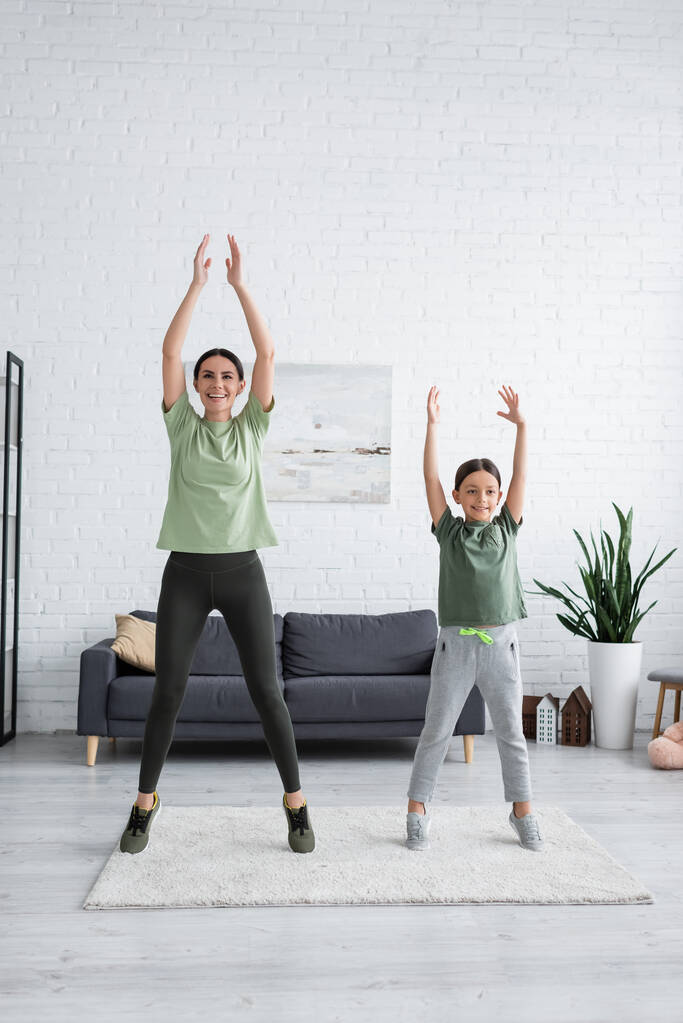 happy girl and nanny stretching on tiptoes with raised hands in living room - Foto, immagini