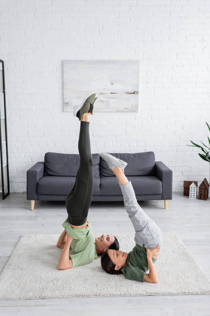happy girl with babysitter training in supported shoulder stand pose on carpet in living room - Photo, Image