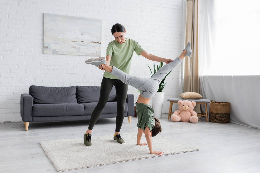 smiling babysitter supporting girl doing handstand in living room - Foto, Imagem