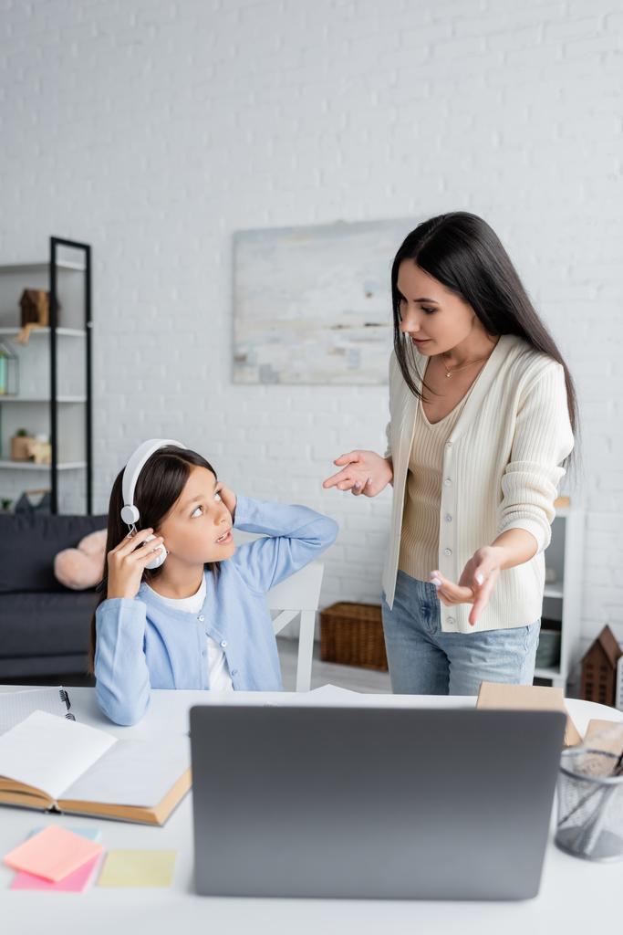 nanny pointing at laptop near girl sitting in headphones during online lesson at home - Photo, image