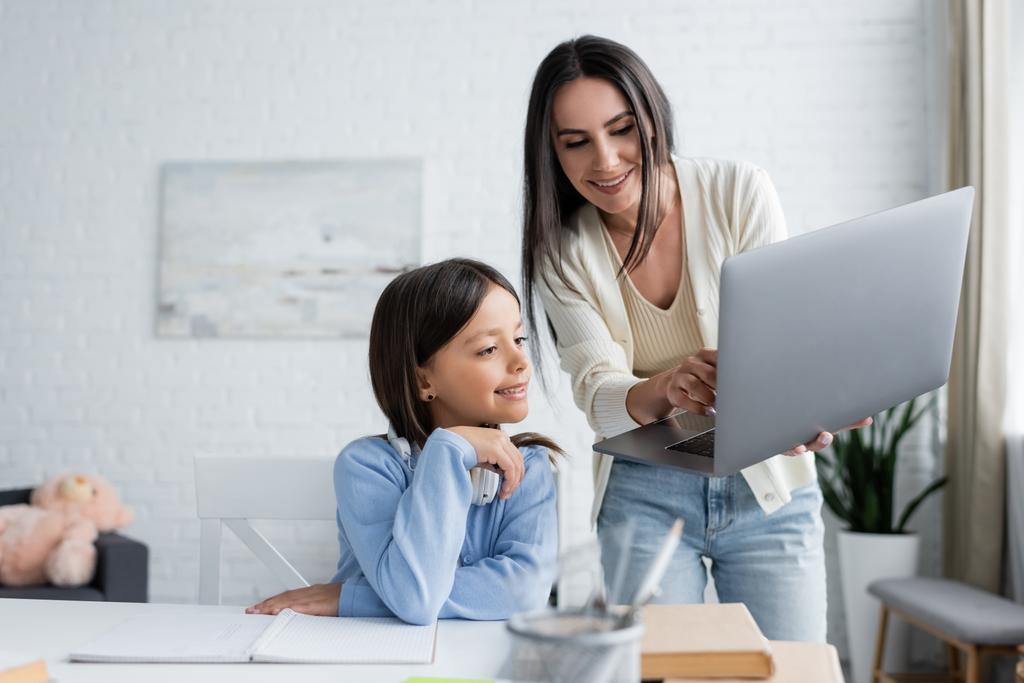 babysitter showing laptop to smiling girl doing homework on blurred foreground - Fotoğraf, Görsel