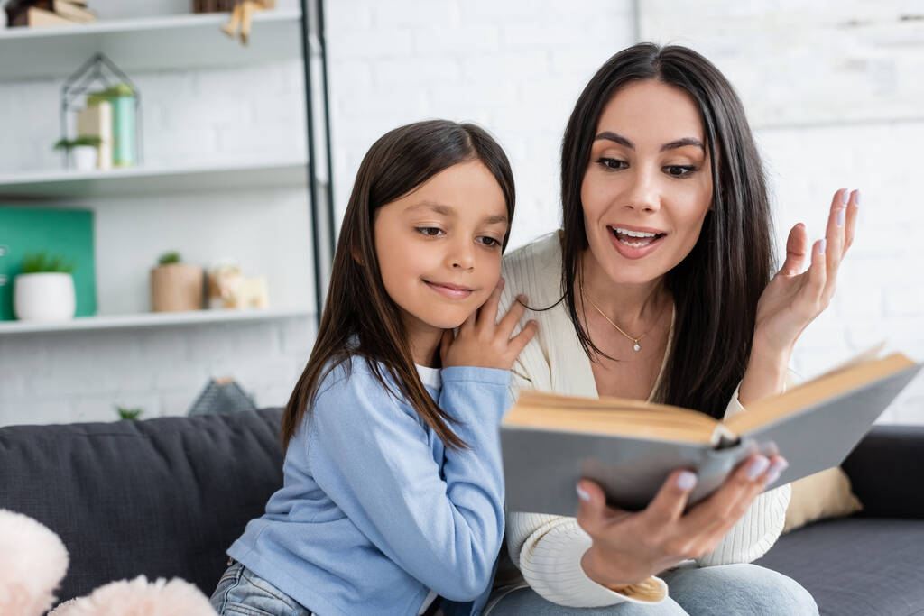 babysitter reading book to smiling girl and gesturing on blurred foreground - Photo, Image