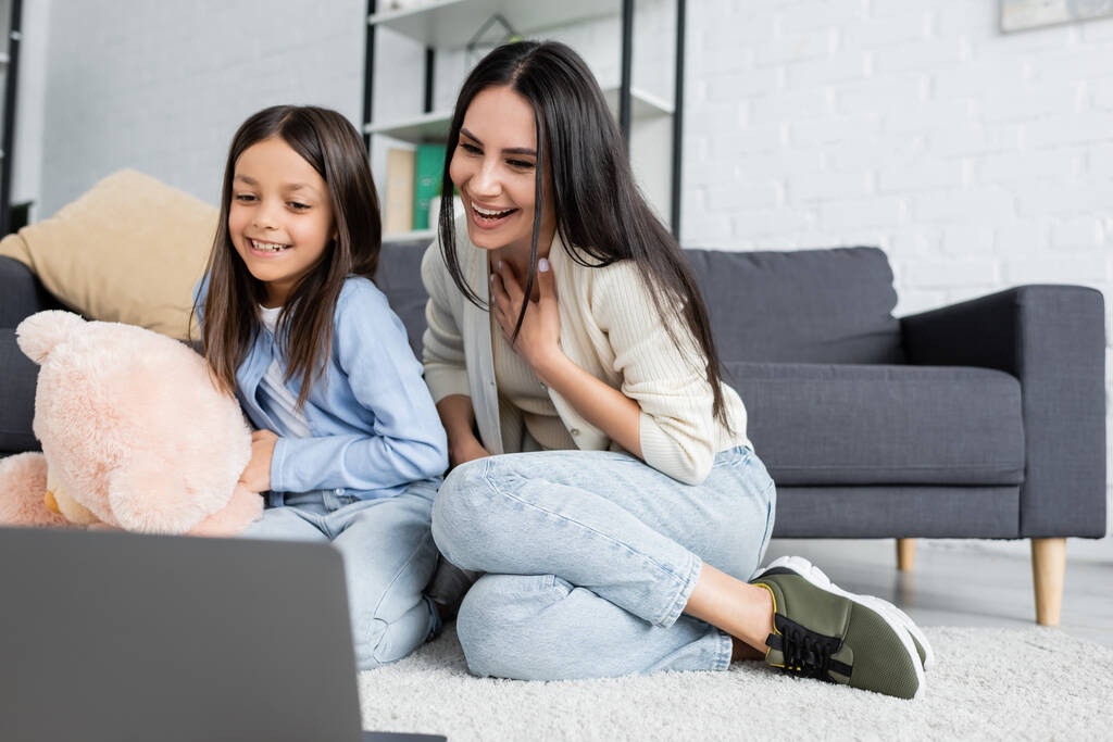 cheerful girl and nanny watching movie on laptop while sitting on floor - Foto, Imagen