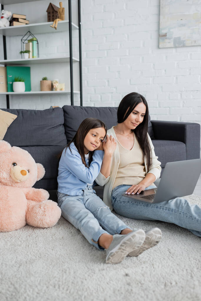 girl leaning on nanny while watching movie on laptop near teddy bear on floor - Photo, Image