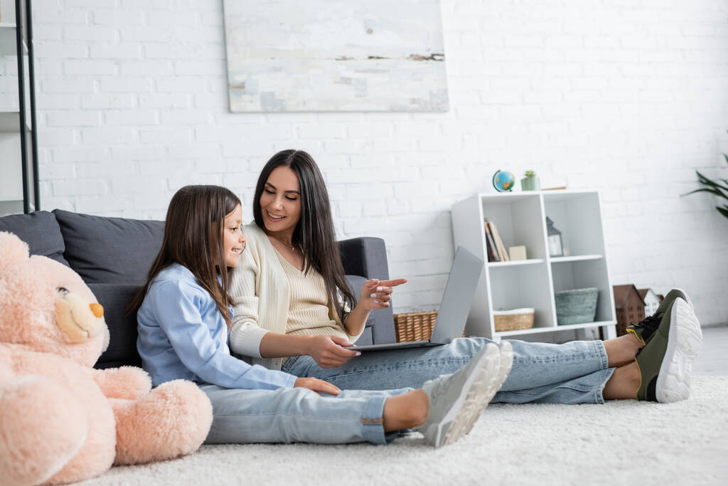 nanny pointing at laptop while watching movie with girl on floor in living room - Photo, image