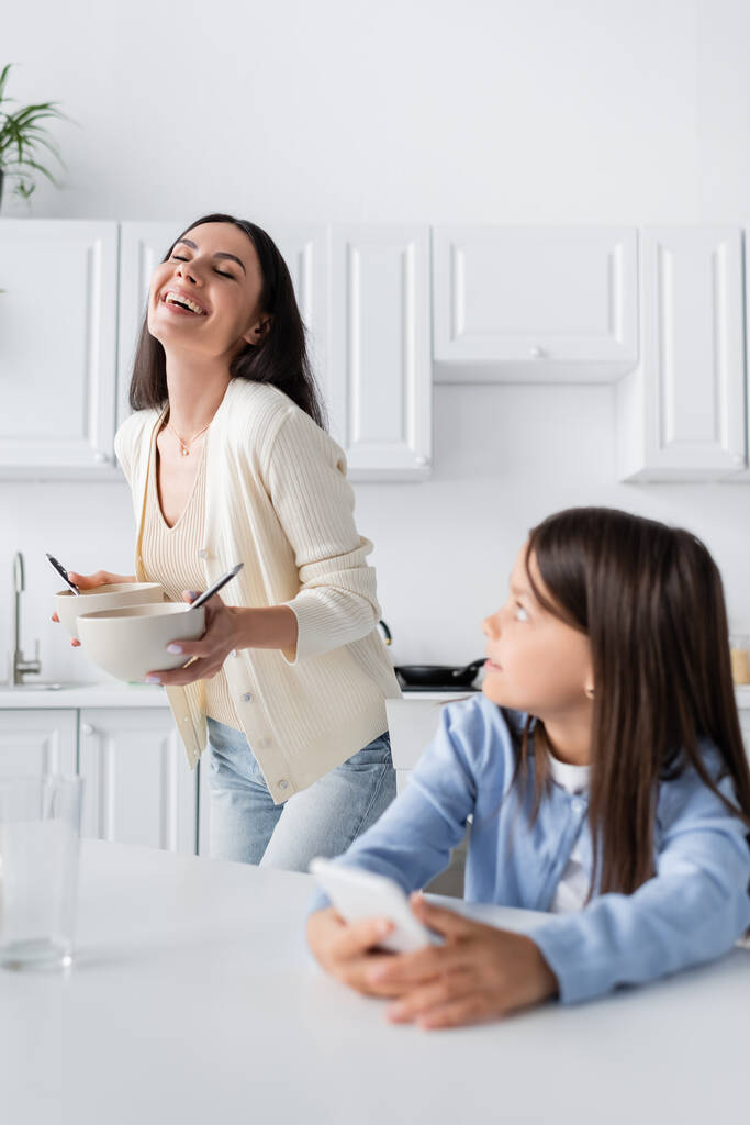 excited babysitter holding bowls with breakfast and laughing near girl in kitchen - Foto, imagen
