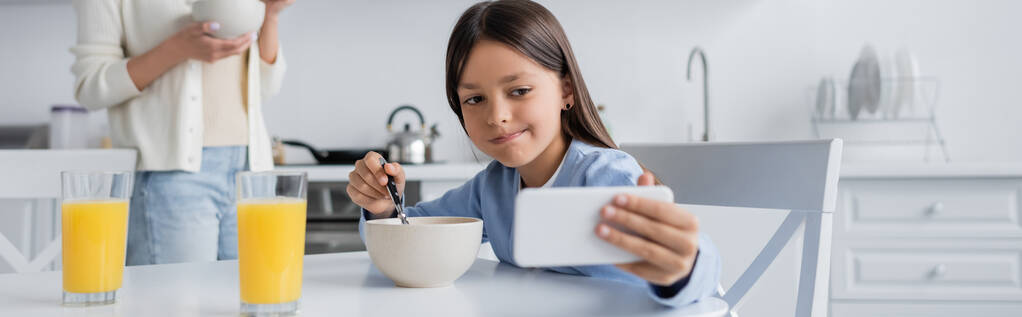 brunette girl grimacing while taking selfie on mobile phone during breakfast near blurred babysitter, banner - Photo, image