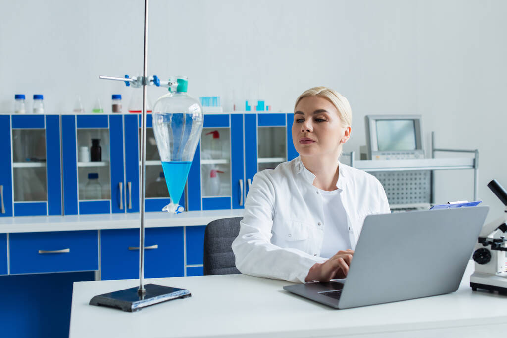 Scientist using laptop and looking at flask with liquid in laboratory  - Foto, imagen