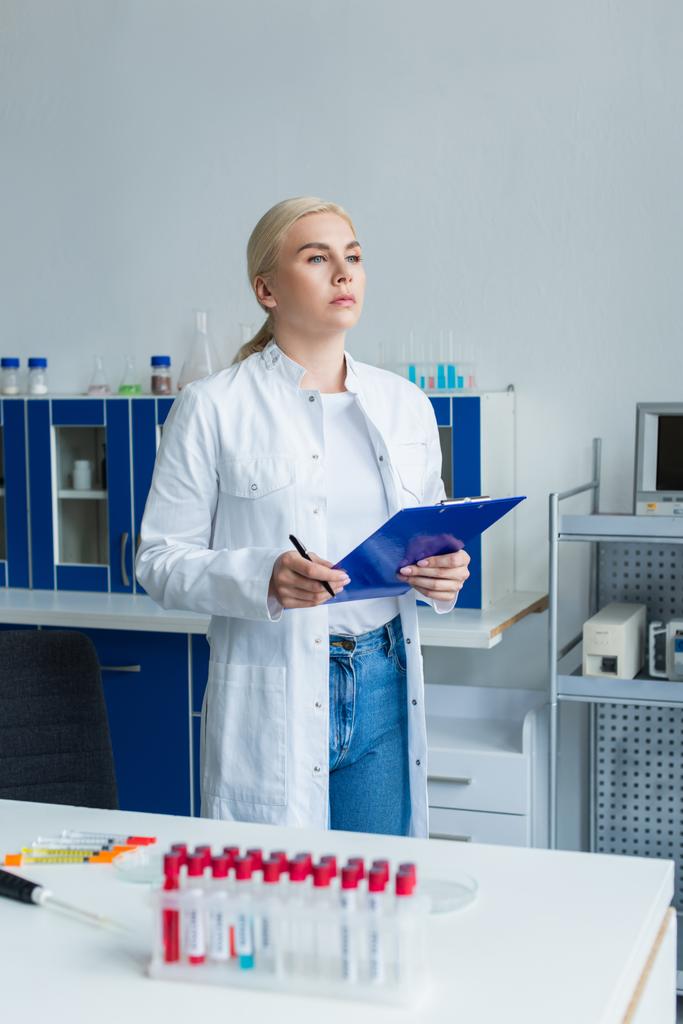 Wetenschapper in witte jas houdt klembord in de buurt van reageerbuizen en elektronische pipet in het lab - Foto, afbeelding