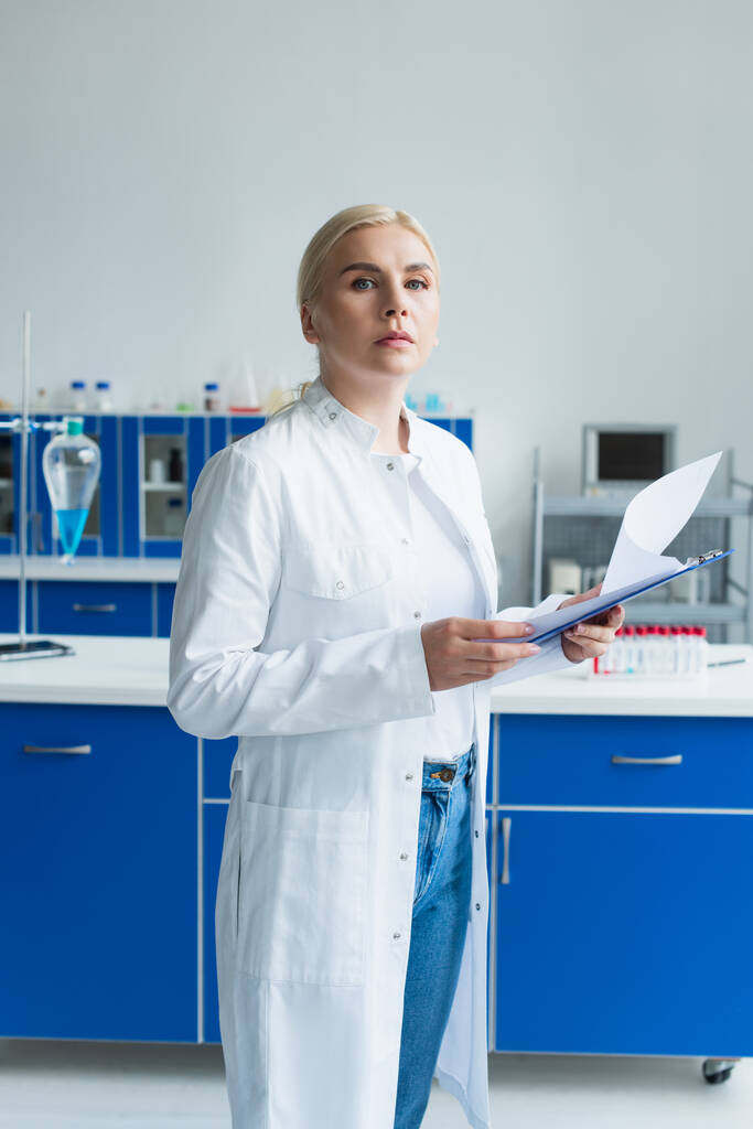 Scientist in white coat holding clipboard and looking at camera in lab  - Foto, Imagen