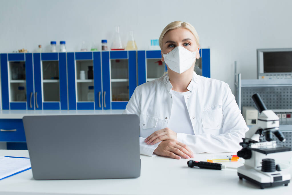 Ученый в защитной маске смотрит в камеру возле микроскопа и ноутбука в лаборатории  - Фото, изображение