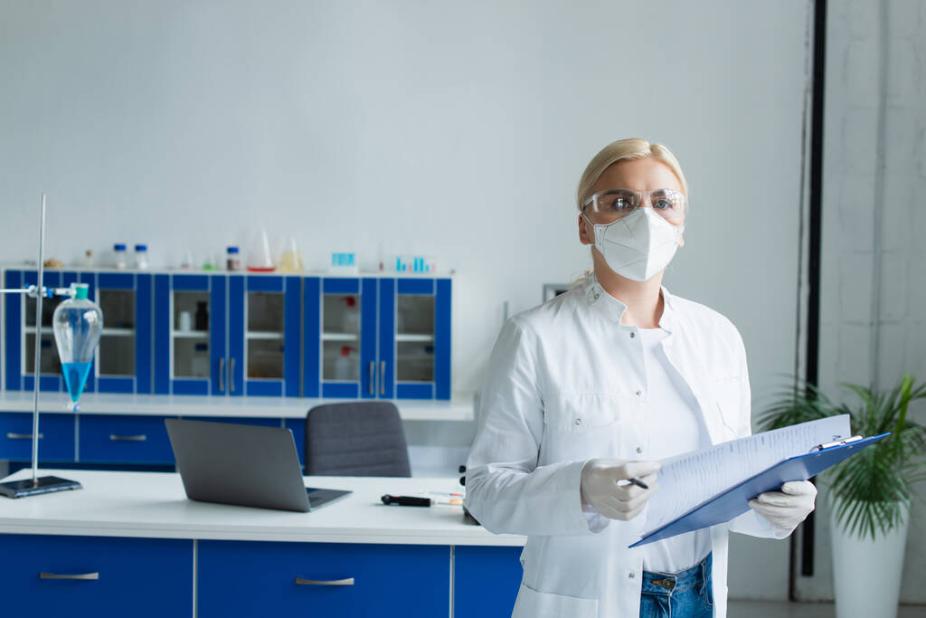 Scientist in googles and protective mask holding clipboard and looking at camera in lab - Fotó, kép
