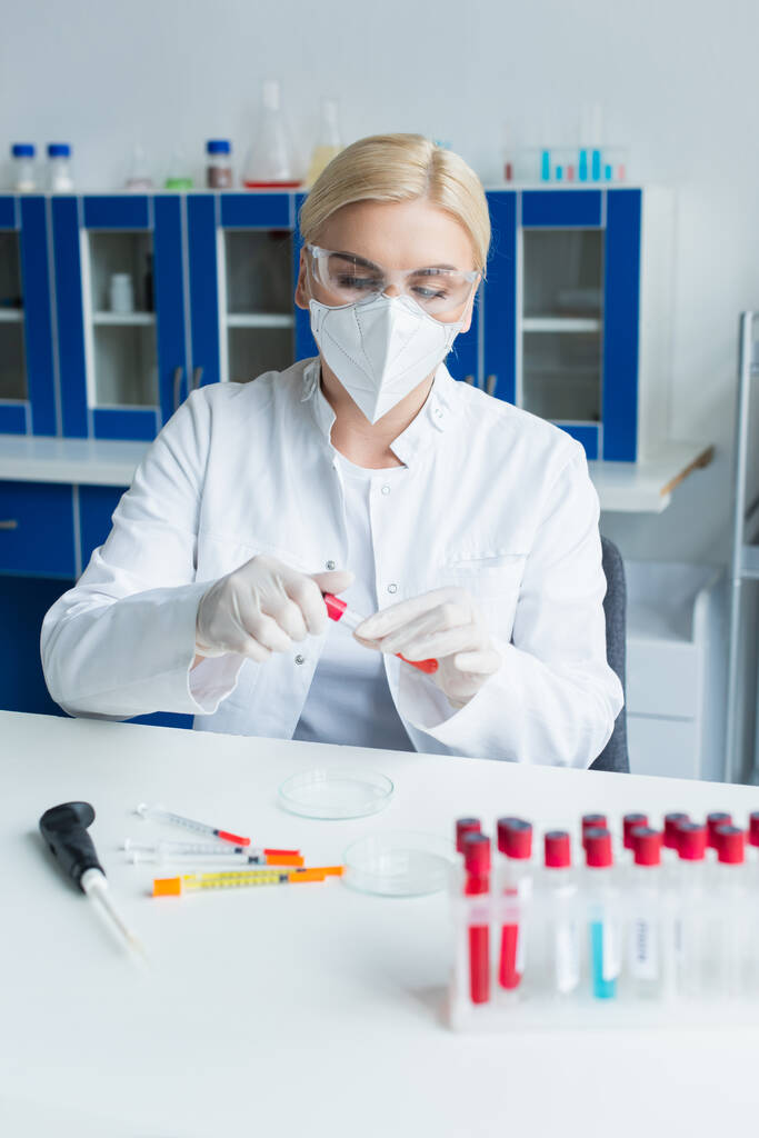 Blonde scientist in protective mask holding test tube near syringes in laboratory - Fotó, kép