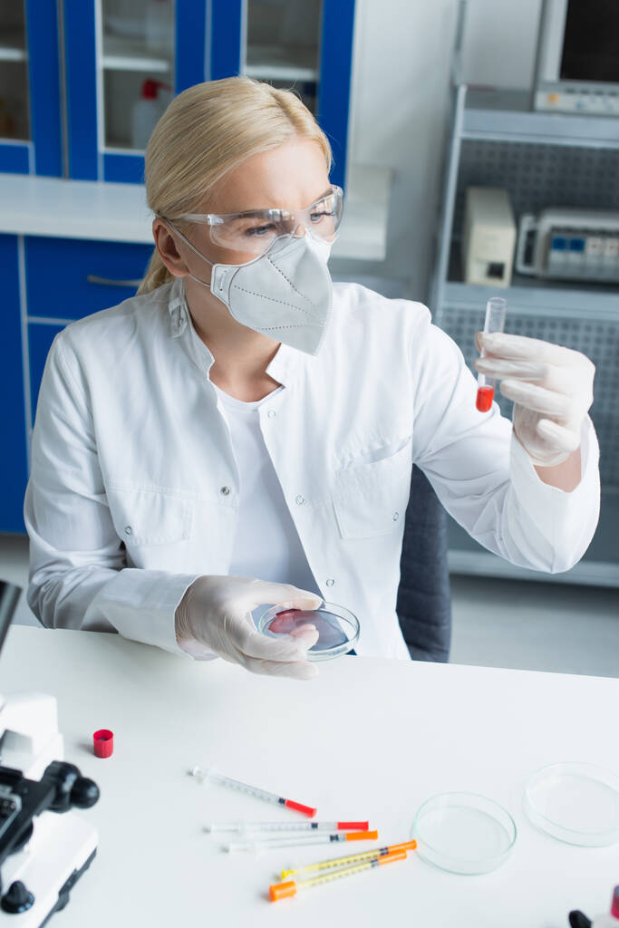 Учёный в очках и маске держит пробирку и чашку Петри рядом со шприцами в лаборатории  - Фото, изображение
