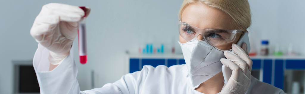 Scientist in protective mask talking on smartphone and holding blurred test tube in lab, banner  - Foto, Imagen