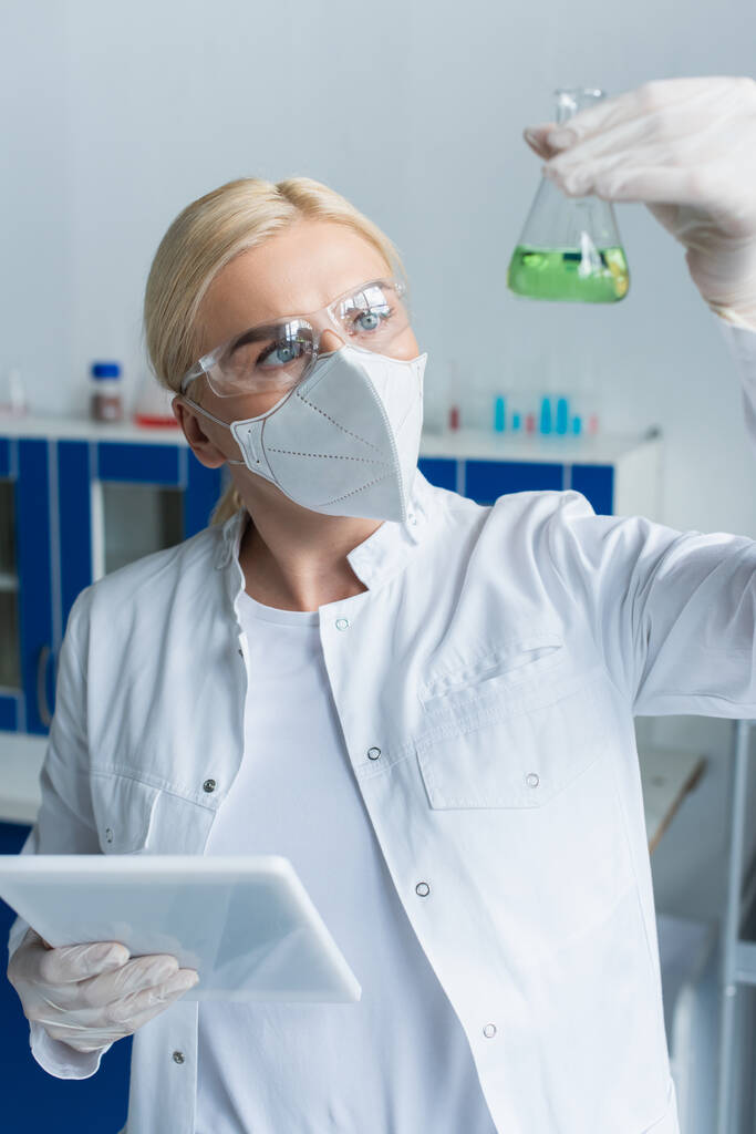 Scientist in goggles looking at blurred flask and holding digital tablet in lab  - Foto, Imagen
