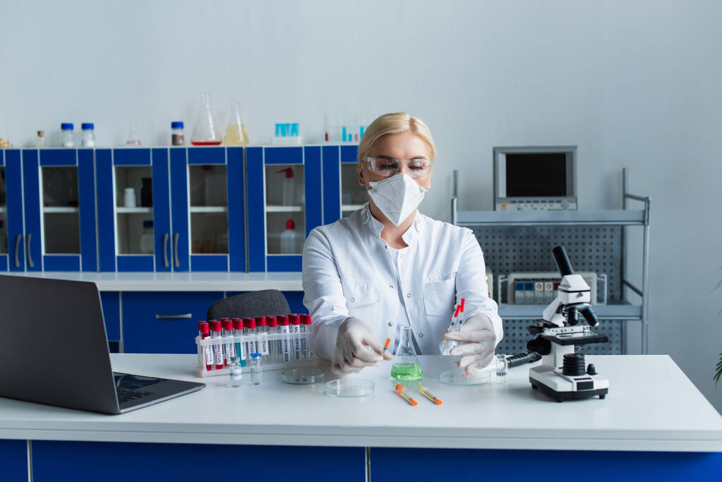 Scientist in protective mask holding syringes near flask and test tubes in lab  - Zdjęcie, obraz