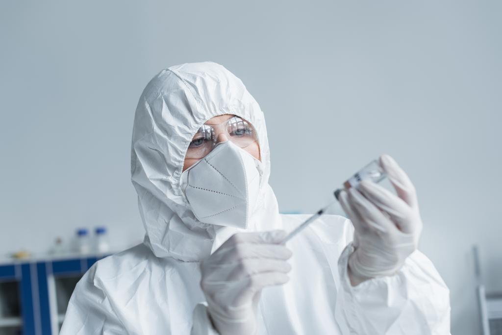 Scientist in protective suit and goggles holding syringe and vaccine in lab  - Photo, Image