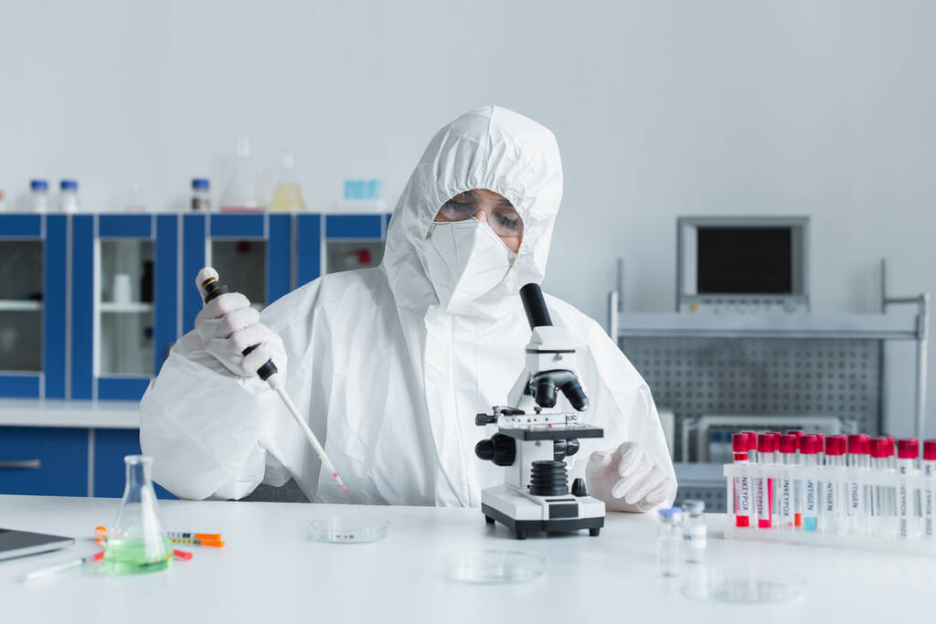 Scientist in protective suit holding electronic pipette near microscope and test tubes in lab  - Fotó, kép
