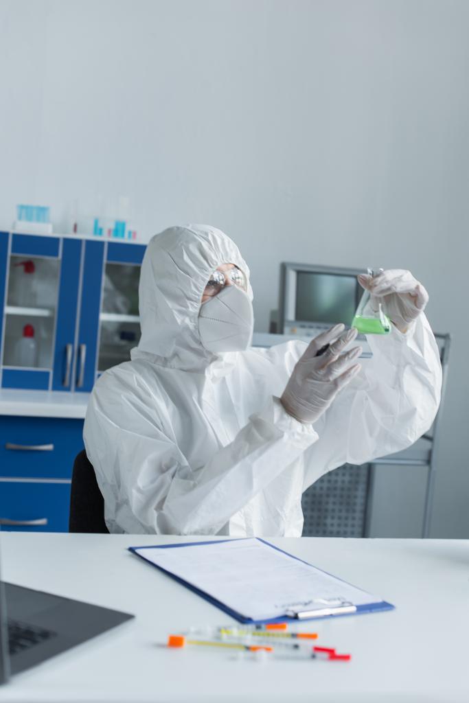 Scientist in hazmat suit holding flask near blurred clipboard and laptop in laboratory  - 写真・画像