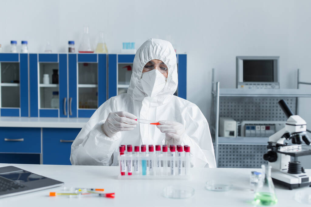 Wissenschaftler mit Schutzbrille und Anzug halten Reagenzglas in der Nähe von Laptop und Mikroskop im Labor  - Foto, Bild