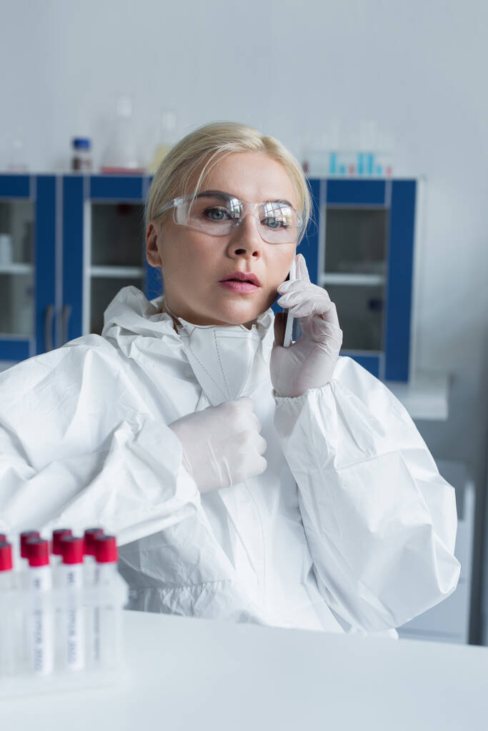 Koruyucu kıyafetli bir bilim adamı laboratuardaki bulanık test tüplerinin yanında akıllı telefondan konuşuyor.  - Fotoğraf, Görsel