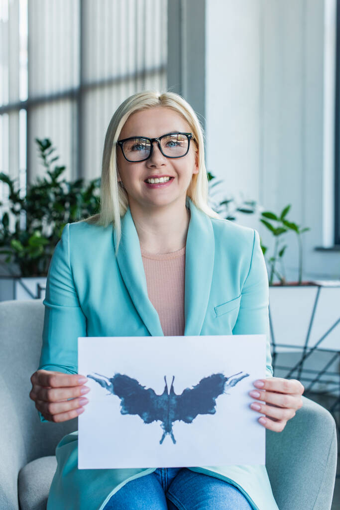 Smiling psychologist in eyeglasses holding Rorschach test in consulting room  - Fotó, kép