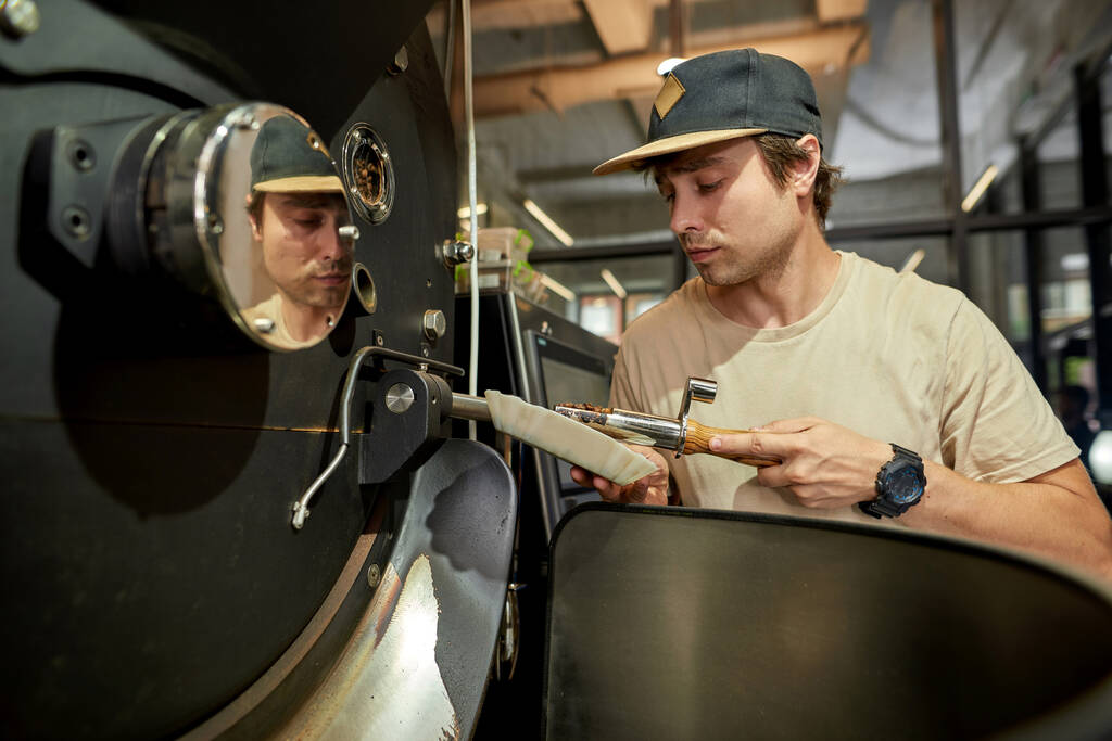 Férfi munkás vagy üzlettulajdonos ellenőrzi kávébab pörkölő gép munka közben a gyárban. Kávékészítés és -gyártás. Kisvállalkozás. Modern gyártó berendezések. Fiatal európai férfi - Fotó, kép