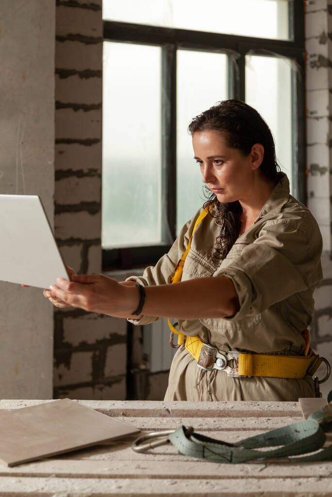 красива жінка в сірому комбінезоні стоїть на будівельнику і тримає в руках відкритий ноутбук. Жіночий портрет без ретуші з її природною недосконалістю
 - Фото, зображення