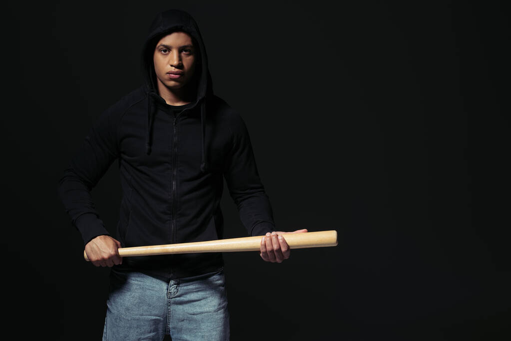 Afrikai amerikai férfi kapucnis pulóverben baseball ütővel a kezében, és a fekete kamerát nézi.  - Fotó, kép