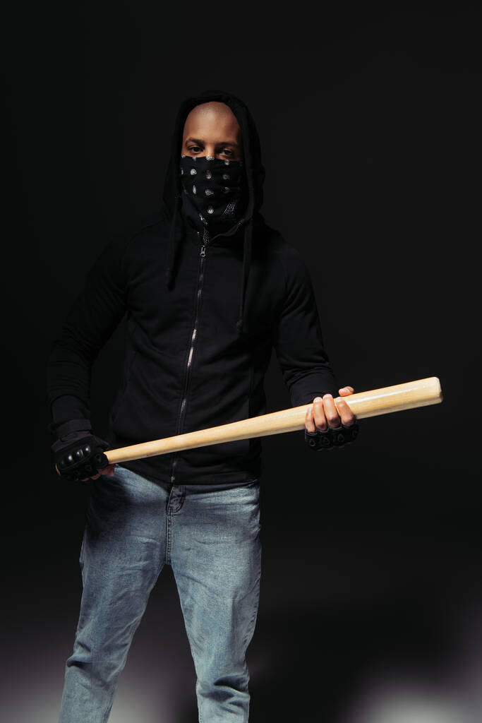 African american hooligan with covered face holding baseball bat on black background  - Fotografie, Obrázek
