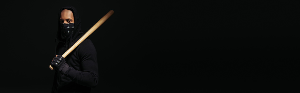 Африканский американский хулиган с маской на лице, держащий бейсбольную биту изолированной на черном, баннер  - Фото, изображение